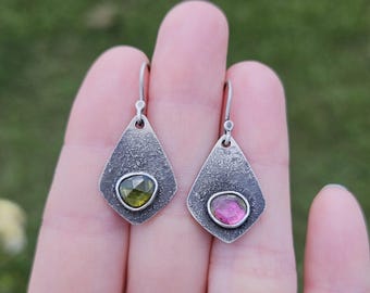May include: A pair of silver earrings with a textured finish. Each earring features a teardrop shape with a bezel-set gemstone. One earring has a green gemstone, the other a pink gemstone. The earrings are displayed against a blurred green background.