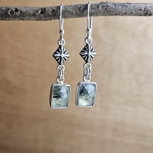 May include: A pair of silver earrings with a geometric design. Each earring features a square, light green gemstone set in a silver frame, suspended from a decorative silver element. The earrings are displayed on a wooden branch.