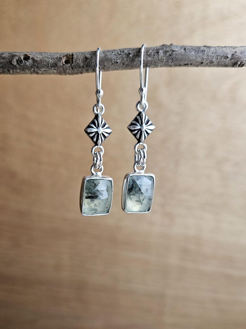 May include: A pair of silver earrings with a geometric design. Each earring features a square, light green gemstone set in a silver frame, suspended from a decorative silver element. The earrings are displayed on a wooden branch.