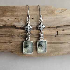 May include: A pair of silver earrings with a rectangular, light green stone. The earrings have a decorative silver element above the stone and a hook for the ear. The earrings are displayed on a piece of wood.