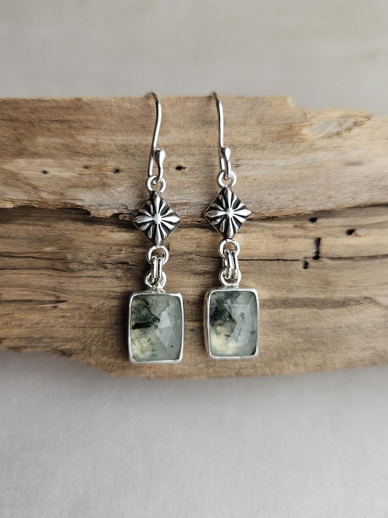 May include: A pair of silver earrings with a rectangular, light green stone. The earrings have a decorative silver element above the stone and a hook for the ear. The earrings are displayed on a piece of wood.