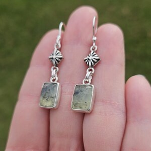 May include: A pair of silver dangle earrings. Each earring features a rectangular, light green gemstone set in a silver bezel. Above the gemstone is a decorative silver element, and the earrings hang from silver hooks.