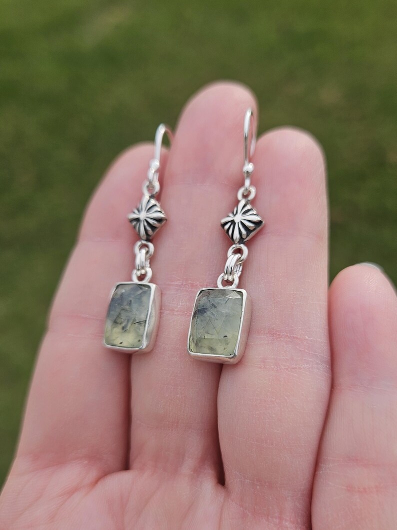 May include: A pair of silver dangle earrings. Each earring features a rectangular, light green gemstone set in a silver bezel. Above the gemstone is a decorative silver element, and the earrings hang from silver hooks.