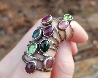 May include: A set of nine silver rings with oval-shaped gemstones. The rings feature a hammered texture and are stacked on a finger. The gemstones are various shades of green and pink.