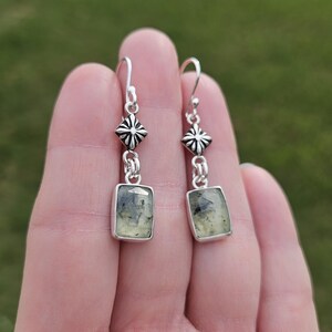 May include: A pair of silver earrings with a rectangular, light green gemstone. The earrings have a decorative silver design above the gemstone and a hook closure. The background is a blurred green.