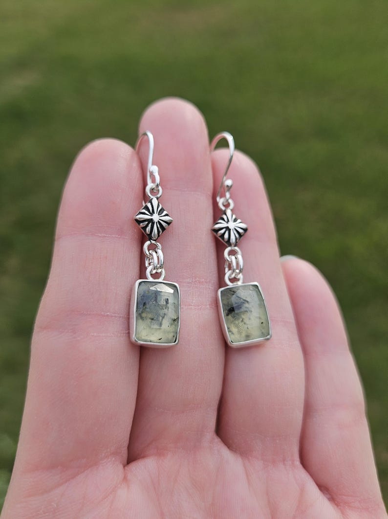 May include: A pair of silver earrings with a rectangular, light green gemstone. The earrings have a decorative silver design above the gemstone and a hook closure. The background is a blurred green.