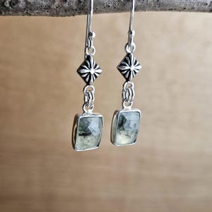 May include: A pair of silver earrings with a geometric design. Each earring features a square, light green gemstone suspended from a decorative silver element. The earrings are hanging from a branch against a blurred background.