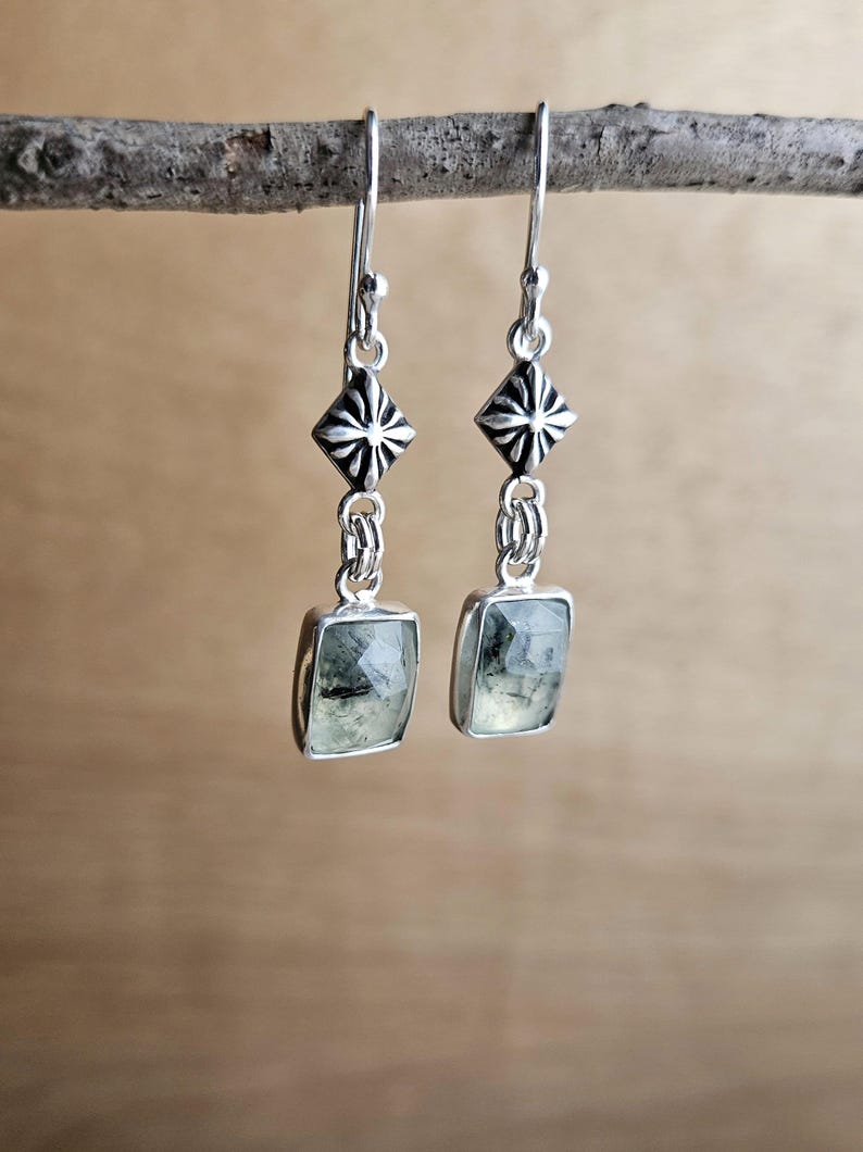 May include: A pair of silver earrings with a geometric design. Each earring features a square, light green gemstone suspended from a decorative silver element. The earrings are hanging from a branch against a blurred background.