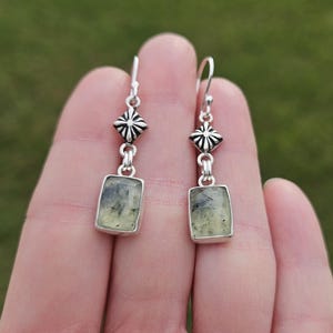May include: A pair of silver earrings with a geometric design. Each earring features a rectangular, light green stone with dark inclusions, set in a silver frame. The earrings have a decorative silver top with a starburst design.