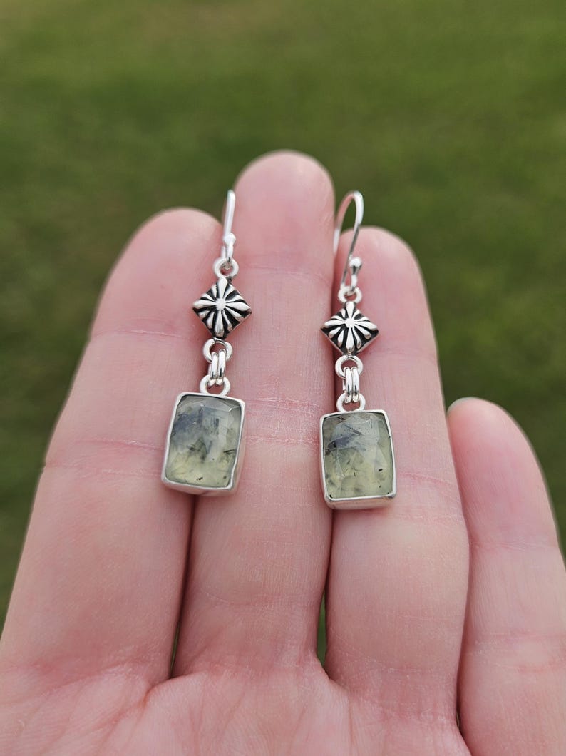 May include: A pair of silver earrings with a geometric design. Each earring features a rectangular, light green stone with dark inclusions, set in a silver frame. The earrings have a decorative silver top with a starburst design.