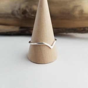 May include: A minimalist silver ring with a V-shaped design. The ring is displayed on a light brown wooden cone, with a blurred wooden background. The ring's simple, elegant design makes it a versatile accessory.