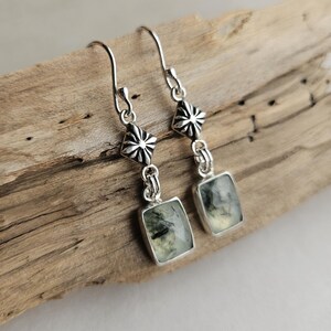 May include: A pair of silver earrings with a rectangular green gemstone pendant. The earrings have a decorative floral design at the top and are displayed on a piece of driftwood.