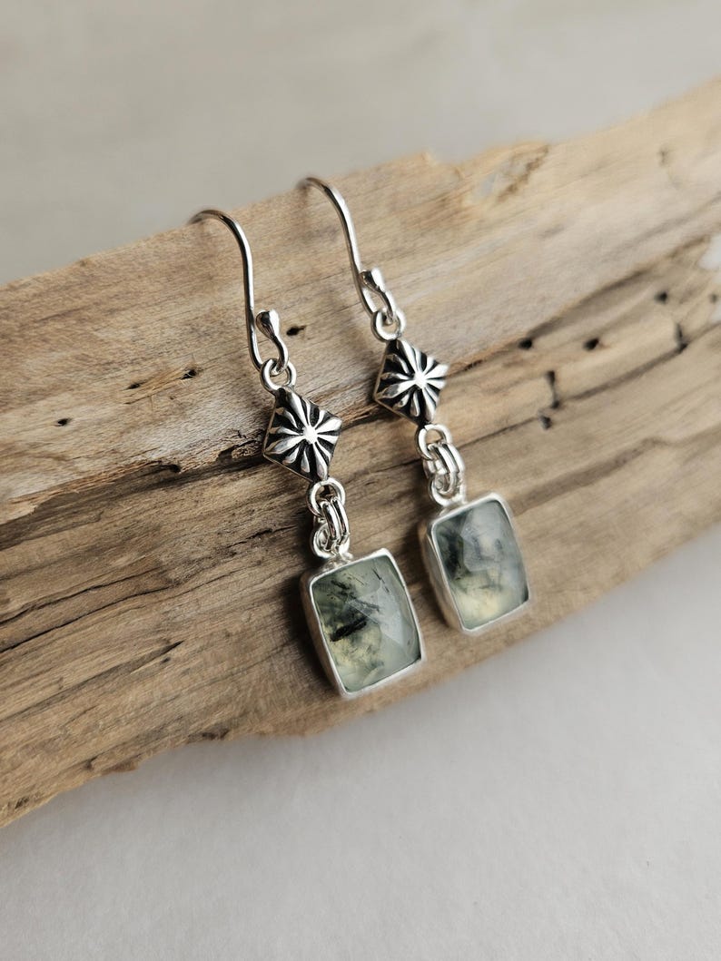 May include: A pair of silver earrings with a rectangular green gemstone pendant. The earrings have a decorative floral design at the top and are displayed on a piece of driftwood.