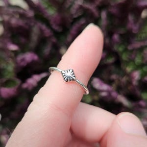 May include: A silver ring with a square, floral-like design. The ring is worn on a finger, with a blurred background of purple foliage. The ring's design is centered on the finger, showcasing its intricate details.