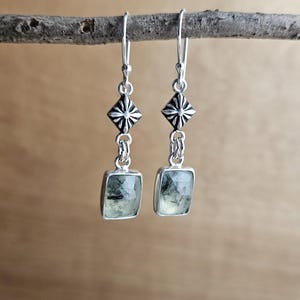 May include: A pair of silver earrings with a geometric design. Each earring features a square, light green stone with dark inclusions, suspended from a decorative silver element. The earrings are displayed on a natural wooden branch.