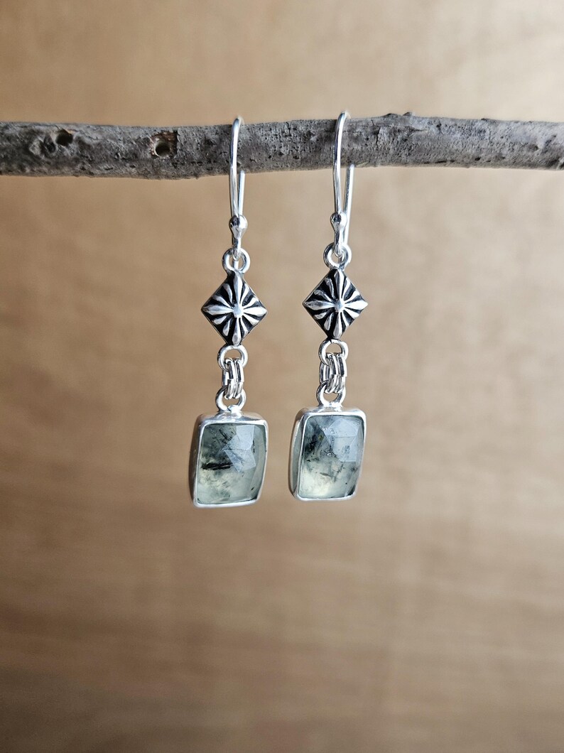May include: A pair of silver earrings with a geometric design. Each earring features a square, light green stone with dark inclusions, suspended from a decorative silver element. The earrings are displayed on a natural wooden branch.