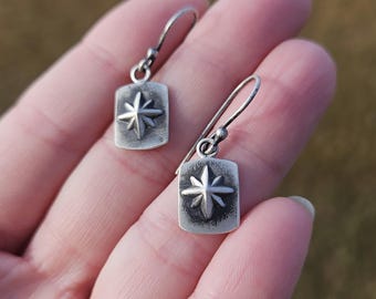 May include: A pair of silver earrings. Each earring features a rectangular pendant with a raised starburst design. The earrings have a simple hook closure. The metal has a slightly aged, textured appearance.