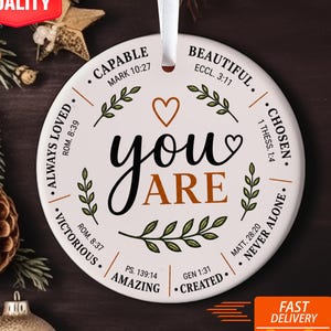May include: A round white ceramic ornament hanging from a white ribbon. The center features the text "YOU ARE" in black and orange, surrounded by phrases like "CAPABLE," "BEAUTIFUL," "CHOSEN," "NEVER ALONE," "CREATED," "AMAZING," "VICTORIOUS," and "ALWAYS LOVED."