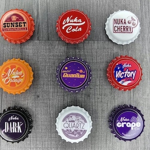 May include: Nine bottle caps with different designs and text. The caps feature the phrases "Sunset Sarsaparilla," "Nuka Cola," "Nuka Cherry," "Nuka Orange," "Quantum," "Nuka Victory," "Nuka Dark," "Quartz," and "Nuka Grape Cola."