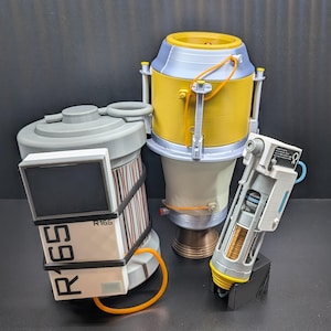 May include: Three-dimensional models of futuristic-looking devices. One is cylindrical with a screen and the text "R165". Another is a yellow and silver container. The third is a white and gray device with a black stand.