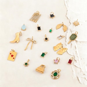 Puede incluir: Una colección de colgantes dorados en varias formas, incluyendo una bota de vaquero, un lazo, una mariposa y una raqueta de tenis. Algunos colgantes presentan piedras preciosas en colores como verde, rosa y rojo. Los colgantes son para joyería.