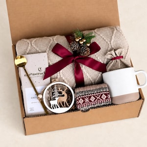May include: A gift box containing a beige cable-knit blanket tied with a burgundy ribbon, a Moonstruck chocolate bar, a gold spoon, a white and tan mug, a small patterned pouch, a pair of patterned socks, and a wooden deer ornament.