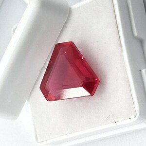 May include: A faceted, triangular-shaped gemstone with a deep red hue. The gem is set in a white, rectangular case with a hinged lid. The gem's cut allows light to reflect, creating a brilliant appearance. The overall presentation suggests a precious stone.