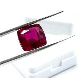 May include: A cushion-cut, deep red ruby gemstone is held by tweezers against a white background. The ruby has a vibrant, saturated color and a polished surface, showcasing its facets. The tweezers are silver-colored.