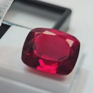 Può includere: Una pietra preziosa taglio a cuscino di un colore rosso intenso e vibrante, con superfici sfaccettate che riflettono la luce. La pietra è posizionata su una base espositiva bianca con cornice nera, evidenziandone la traslucenza e la forma.