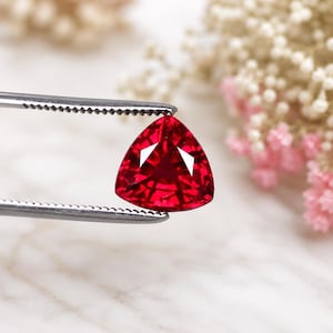 May include: A vibrant, deep red gemstone cut into a triangular shape with multiple facets that reflect light. The stone is held securely by a pair of silver-toned metal tweezers, showcasing its brilliant color and precise geometric cut clearly.