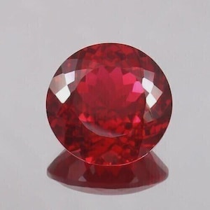 Pode incluir: Uma pedra preciosa redonda e facetada com uma cor vermelha profunda e vibrante. A pedra apresenta um corte preciso e simétrico com múltiplas facetas triangulares e em forma de pipa que refletem a luz, criando um aspeto brilhante e cintilante.