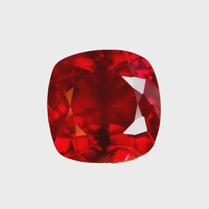 May include: A cushion-cut, faceted gemstone with a deep red hue. The gem's surface reflects light, showcasing its internal structure and brilliance. The shape is a square with rounded corners, typical of cushion cuts.