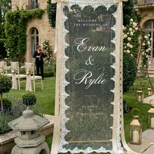 May include: A transparent wedding sign with white lace edging and cream ribbons. The sign displays "Welcome to the Wedding of Evan & Rylie" and the date February 1, 2026. The sign is set up in a garden, with a violinist in the background.