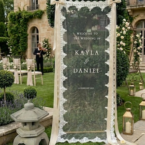 May include: A transparent wedding sign with a decorative white lace border and cream ribbons. The sign displays "Welcome to the Wedding of Kayla & Daniel, 14 February 2025." The sign is placed on a green lawn, with a stone lantern and a violinist visible.