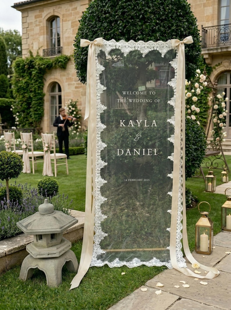 May include: A transparent wedding sign with a decorative white lace border and cream ribbons. The sign displays "Welcome to the Wedding of Kayla & Daniel, 14 February 2025." The sign is placed on a green lawn, with a stone lantern and a violinist visible.
