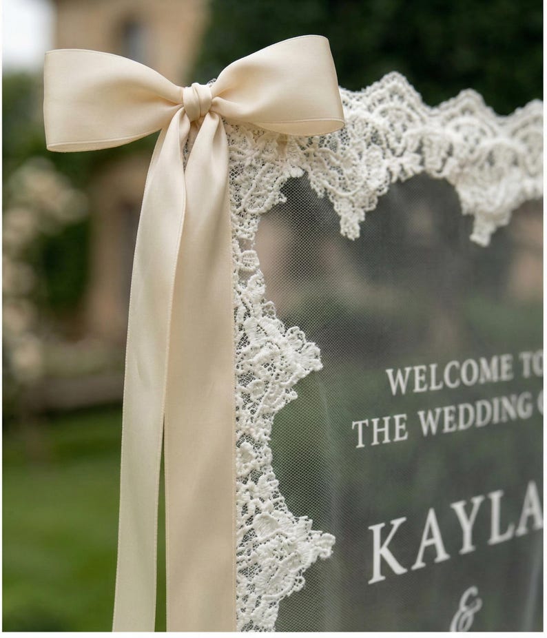 May include: A wedding welcome sign featuring a cream satin bow and ribbon. The sign is trimmed with white lace and displays the words "WELCOME TO THE WEDDING KAYLA &" in white.