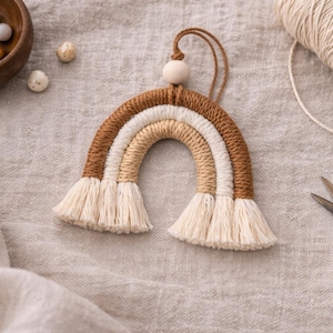 May include: A handcrafted macramé rainbow in brown, cream, and beige, with a wooden bead and hanging loop. A wooden bowl with decorative balls, a spool of twine, and scissors are also in the image. The rainbow is a decorative item.