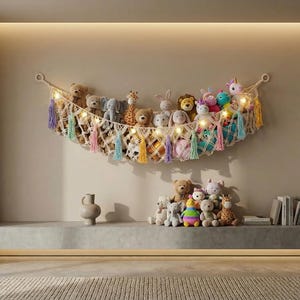 Könnte beinhalten: Eine Makramee-Hängematte mit Stofftieren wie Teddybären, einer Giraffe und einem Einhorn hängt an einer Wand. Die Hängematte ist mit bunten Quasten und Lichterketten verziert. Zusätzliche Stofftiere befinden sich auf einem Regal darunter.