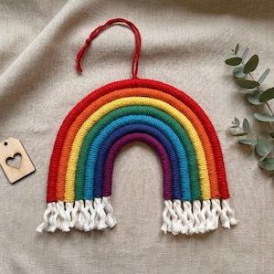 Może przedstawiać: Ręcznie robiony tęczowy wieszak ścienny z czerwoną pętlą do zawieszania. Tęcza ma żywe kolory, w tym czerwony, pomarańczowy, żółty, zielony, niebieski i fioletowy. Z dołu zwisają białe frędzle. Obecna jest również mała drewniana etykieta z wyciętym sercem.