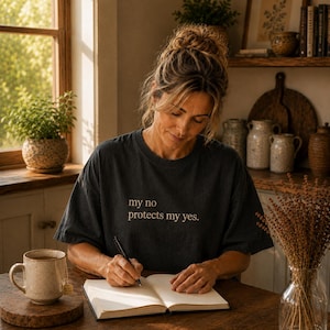 Könnte beinhalten: Eine Frau trägt ein dunkelgraues T-Shirt mit dem Text "my no protects my yes." Sie sitzt an einem Holztisch und schreibt in ein Notizbuch. Eine Tasse Tee und eine Vase mit Trockenblumen stehen ebenfalls auf dem Tisch.