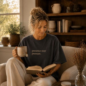 Könnte beinhalten: Ein dunkelgraues T-Shirt mit dem Text "presence over pressure" in heller Farbe. Die Person hält eine Tasse und liest ein Buch. Die Szene spielt in einem gemütlichen Wohnzimmer mit Fenster und Bücherregalen.