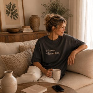 Könnte beinhalten: Ein dunkelgraues T-Shirt mit dem Text "i choose what enters" in heller Farbe. Die Person hält einen beigefarbenen Becher mit gesprenkeltem Design. Die Person sitzt auf einem beigefarbenen Sofa mit dekorativen Kissen.