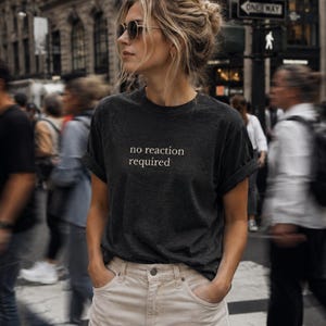 Könnte beinhalten: Dunkelgraues T-Shirt mit dem weißen Text "no reaction required". Die Ärmel sind hochgekrempelt, und das Shirt wird mit hellen Hosen getragen. Der Hintergrund ist unscharf, was auf eine Außenaufnahme hindeutet.