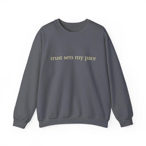 Peut inclure: Un sweat-shirt gris foncé avec l'inscription "trust sets my pace" en or clair. Le sweat-shirt a des manches longues et un col rond. Le tissu semble doux et confortable.