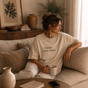 Könnte beinhalten: Eine Person trägt ein beiges T-Shirt mit dem Text "i choose what enters." und sitzt auf einem beigen Sofa. Sie hält eine gesprenkelte Tasse. Auf einem Holztisch liegen ein Buch und ein Smartphone. Dekorative Kissen und eine Vase sind ebenfalls zu sehen.