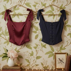 May include: Two corsets hang on gold hangers against a floral wallpaper backdrop. One is a deep burgundy satin with bow straps, the other is navy blue with a lace-up front and bow straps. A peony, books, and a bird print decorate the shelf below.