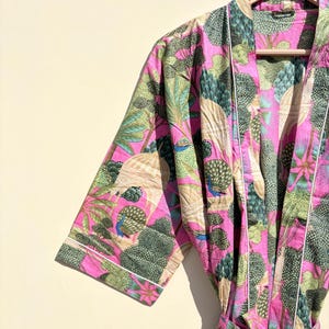 May include: A vibrant pink robe with a botanical print featuring trees, leaves, and peacocks. The robe has white trim and a matching belt. The short-sleeved robe is hanging on a wooden hanger.