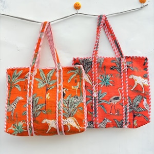 May include: Two orange tote bags with a jungle-themed print featuring zebras, leopards, and palm trees. The bags have striped handles and trim. The bags are hanging against a white wall.