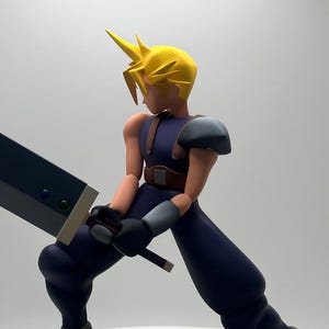 Puede incluir: Figura de un personaje con pelo amarillo puntiagudo, vistiendo un atuendo azul y sosteniendo una gran espada negra. La figura está posada en una postura dinámica sobre una base circular gris, sobre un fondo blanco.