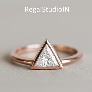 Peut inclure: Une bague en or rose avec une pierre pr&eacute;cieuse blanche triangulaire. La pierre est sertie dans un sertissage &agrave; lunette, et le bracelet est lisse et poli. La bague est pr&eacute;sent&eacute;e sur une surface grise neutre.